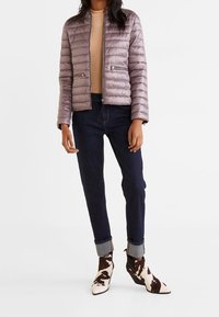 Kvinde iført en lyslilla quiltet jakke, beige sweater, mørke bukser med opsmøg og brune og hvide mønstrede ankelstøvler, der står mod en hvid baggrund.