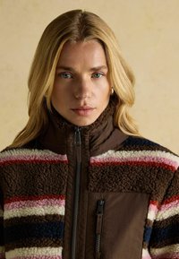 Blonde Frau mit blauen Augen, die eine braune Fleecejacke mit rosa, roten, weißen und marineblauen Streifen sowie Frontreißverschlussdetails trägt.