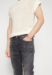 Person wearing a white knitted sleeveless top and faded black jeans with a hand in the front pocket, wearing a silver bracelet.