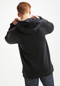 Sweat à capuche noir en matériau doux, avec une grande capuche, une coupe ample et un ourlet côtelé. Porté par une personne vue de dos, avec un jean en denim.