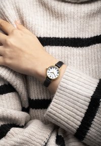 Montre-bracelet ronde avec un cadran blanc, des chiffres romains noirs, un boîtier doré et un bracelet en cuir noir, portée sur une main contre un pull rayé.