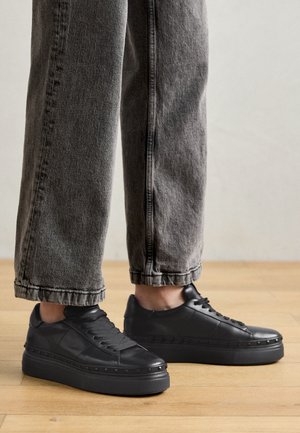 Person iført sorte plateau-sneakers med nittebesætning og brede grå jeans, stående på et trægulv.