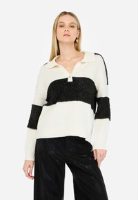 Maglione a righe crema e nere con collo a camicia, caratterizzato da un tessuto nero testurizzato e un dettaglio con bottoni sul collo. Pantaloni neri sotto.