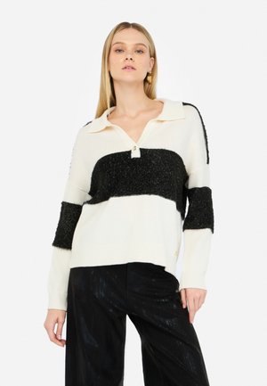 Maglione a righe crema e nere con collo a camicia, caratterizzato da un tessuto nero testurizzato e un dettaglio con bottoni sul collo. Pantaloni neri sotto.