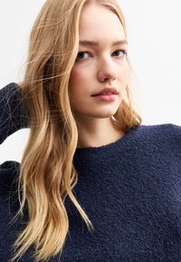 Marinblå, texturerad tröja med rund halsringning, bärs av en person med långt hår. Tyget verkar mjukt och mysigt.