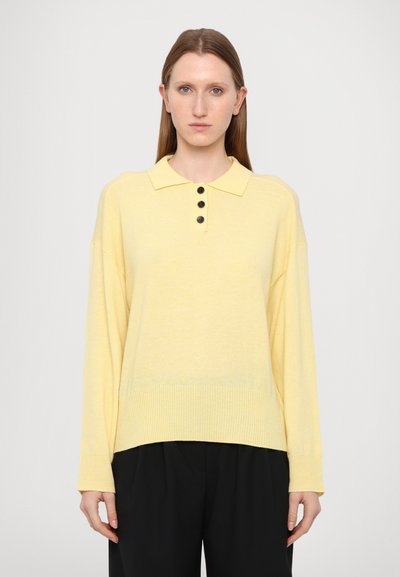 Geel gebreide pullover met een kraag, drie knopen aan de voorkant, lange mouwen en een geribbelde boord; gecombineerd met losse zwarte broeken.