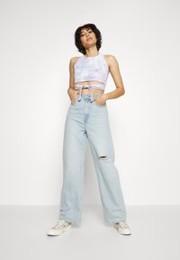 Top cropped em tie-dye roxo com design de halter neck e detalhe em nó, combinado com umas calças de ganga largas em azul claro com joelhos desgastados.
