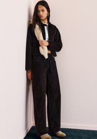 Ruskea corduroy-takki ja siihen sopivat korkeavyötäröiset housut, yhdistettynä valkoiseen paitaan ja beigelle neulotulle huiville, vaaleanpunaisen seinän taustalla.