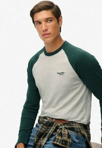Ung mand iført en hvid og mørkegrøn raglanærmer-skjorte med "SuperDry Athletics"-logo, blå jeans og en ternet skjorte bundet omkring sin talje.