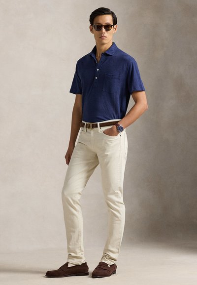 Μπλε ναυτικό κοντομάνικο πόλο με τσέπη στο στήθος, σε συνδυασμό με ελεφαντόδοντο παντελόνια. Το σύνολο ολοκληρώνουν καφέ σουέντ παπούτσια.