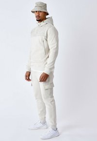 Hoodie beige avec un haut à motifs, poche zippée, et un pantalon de survêtement assorti avec un grand patch. Les accessoires incluent un chapeau seau à motifs et des baskets blanches.