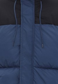 Blaue Pufferjacke mit matter Textur, einem schwarzen oberen Schulterbereich, Kordeln an der Kapuze und gestepptem Futter.