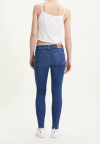 Jeans ajustados de mezclilla de color azul con un dobladillo deshilachado, mostrando un parche de imitación de cuero en la parte trasera de la cintura; modelo vistiendo un crop top blanco.