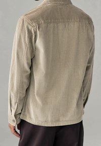 Chemise en velours côtelé beige atténué, avec une texture côtelée verticale, des poignets à boutons et un col classique. Vue de dos montrant les détails des coutures.