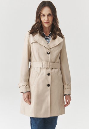 Beige trenchcoat met grote kraag, een ceintuur in de taille en twee voorzakken. Voorzien van donkere knopen en een gladde, lichte stof.