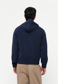 Donkerblauwe lichtgewicht jas met een capuchon, elastische tailleband en raglanmouwen, met een zachte textuur en een minimalistisch ontwerp.