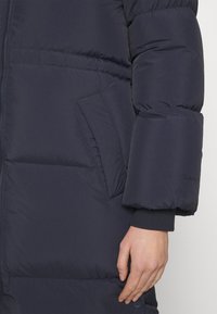 Marineblauer Steppmantel mit Reißverschluss, Seitentasche und gerippten Bündchen. Hergestellt aus glattem, leichtem Stoff mit leicht glänzendem Finish.