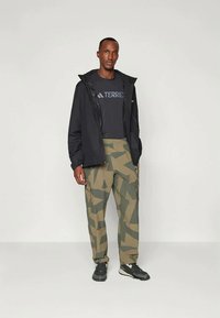 Veste imperméable noire sur un t-shirt gris foncé, accompagnée d'un pantalon cargo camouflage vert. Le modèle porte des chaussures de sport noires, les mains dans les poches.