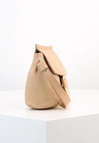 Sac à main en daim beige avec un rabat en cuir lisse, un accent en métal doré et une bandoulière amovible. Forme arrondie avec des détails de couture.