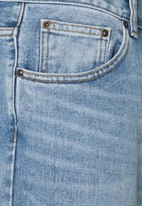 Lichtblauwe denim jeans met een vervaagde afwerking, voorzien van een klein achterzakje met donkere metalen klinknagels en zichtbare stiksels.