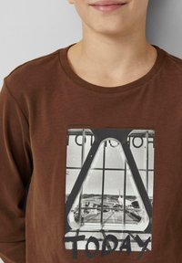 Brązowa koszulka z długim rękawem z dużą monochromatyczną grafiką okna lotniska z napisem „TODAY” w pogrubionych czarnych literach.