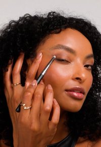 Un pennello per il trucco con manico nero e setole morbide viene utilizzato per applicare il prodotto sulla palpebra. La modella ha capelli ricci e indossa anelli d'oro.