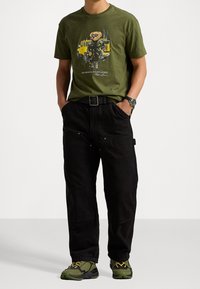 Olivgrön bomullströja med grafisk bild av en tecknad björn, matchad med svarta cargobyxor med fickor och ett svart bälte. Bärs med gröna sneakers.