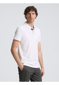 Polo shirt blanc à manches courtes et col, doté de deux boutons de fermeture. Associé à un pantalon texturé gris foncé.