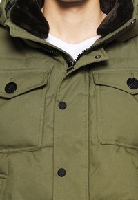 Primo piano di una giacca imbottita verde con bottoni e zip neri, che mostra il collo foderato di pelliccia marrone scuro e una maglietta a girocollo bianca sotto.