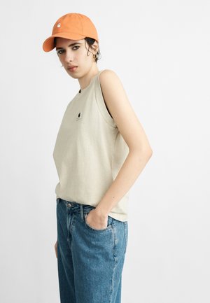 Młoda osoba w pomarańczowej czapce, beżowej koszulce bez rękawów z logo i niebieskich dżinsach, stojąca z jedną ręką w kieszeni na białym tle.