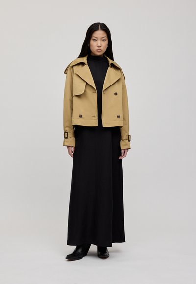 Khaki värviline natuke lühem trench coat suurte krae ja topelt nuppudega, koos vöödetailiga, kantud musta maksikleidiga ja kontsakingadega.