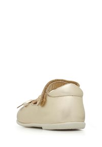 Zapato de cuero dorado con textura suave, forma redondeada y una correa de velcro. Presenta una suela de goma plana y un pequeño lazo decorativo en la parte delantera.