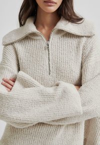 Jasno beżowy sweter na zamek z teksturowanej plecionej tkaniny, szerokim kołnierzem i oversize'owymi rękawami, z metalowym detalem zamka błyskawicznego przy szyi.