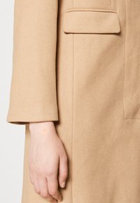 Beige ullkappe med en strukturert design, rette ermer og en enkelt brystlomme. Glatt tekstur med et subtilt vevmønster.
