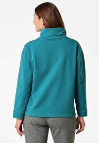 Tegenpullover sweatshirt in teal met een hoge kraag en een ontspannen pasvorm, met lange mouwen en een soepele textuur, gecombineerd met broeken in een houndstooth-patroon.