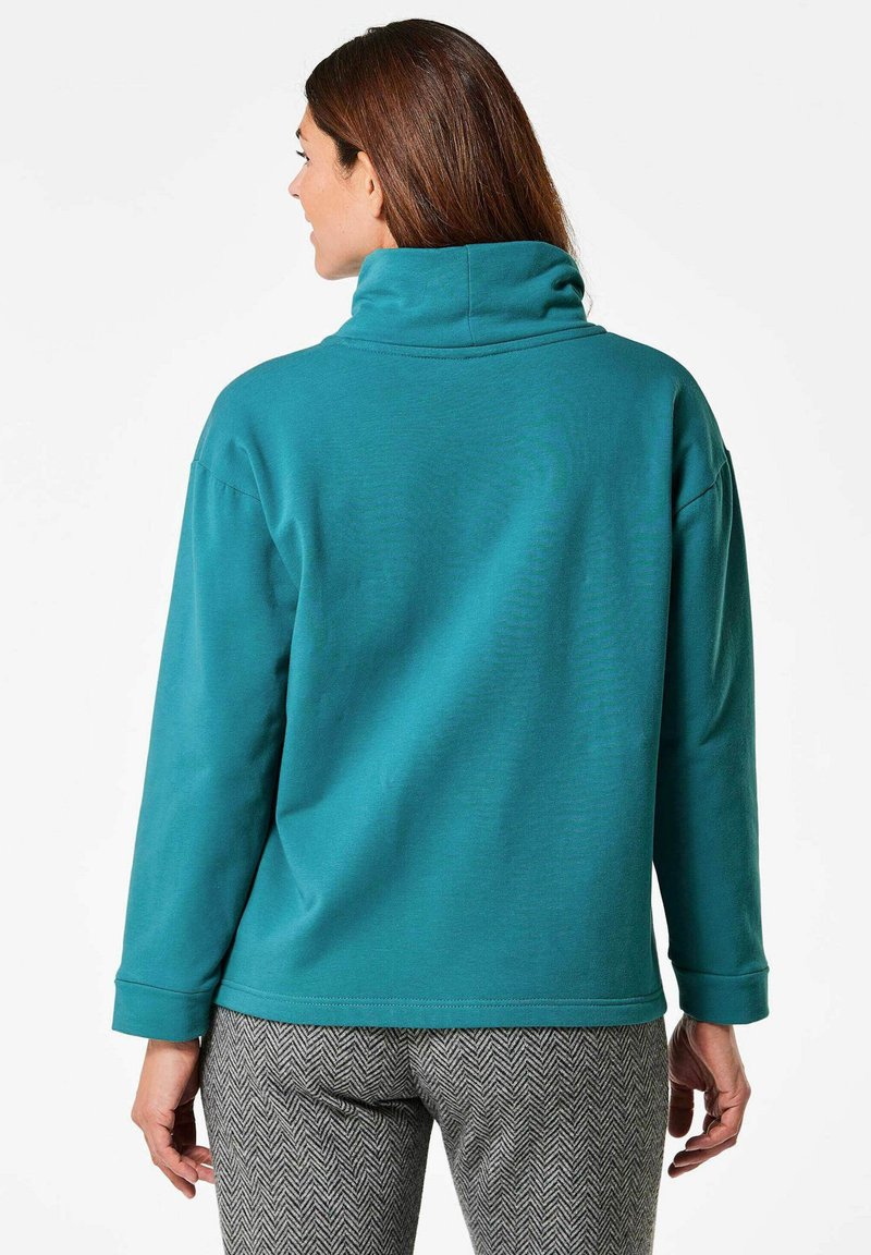 Tegenpullover sweatshirt in teal met een hoge kraag en een ontspannen pasvorm, met lange mouwen en een soepele textuur, gecombineerd met broeken in een houndstooth-patroon.