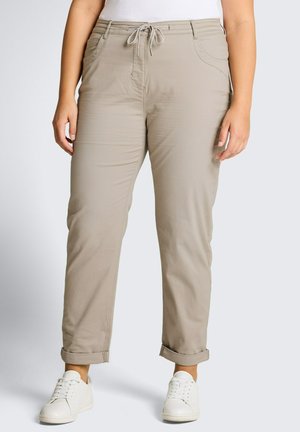 Frau trägt beigefarbene Kordelzughose mit umgeschlagenen Bündchen und weiße Turnschuhe, gezeigt von der Mitte des Torsos bis zu den Füßen vor einem einfachen Hintergrund.