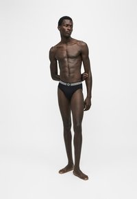 Homme grand et mince à la peau foncée portant un slip noir avec une ceinture à motif, debout pieds nus sur un fond blanc uni.