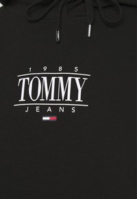 Černá mikina s kapucí, na které je výrazné bílé logo s nápisem "TOMMY JEANS" a datem "1985" nad ním. Obsahuje malý červený, bílý a modrý akcent.