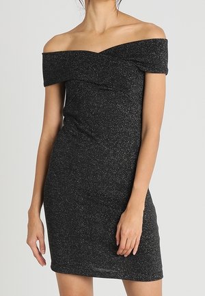 Vrouw die een nauwsluitende, zwartkleurige korte jurk draagt met een subtiele glans en een off-the-shoulder ontwerp, staand tegen een effen achtergrond.