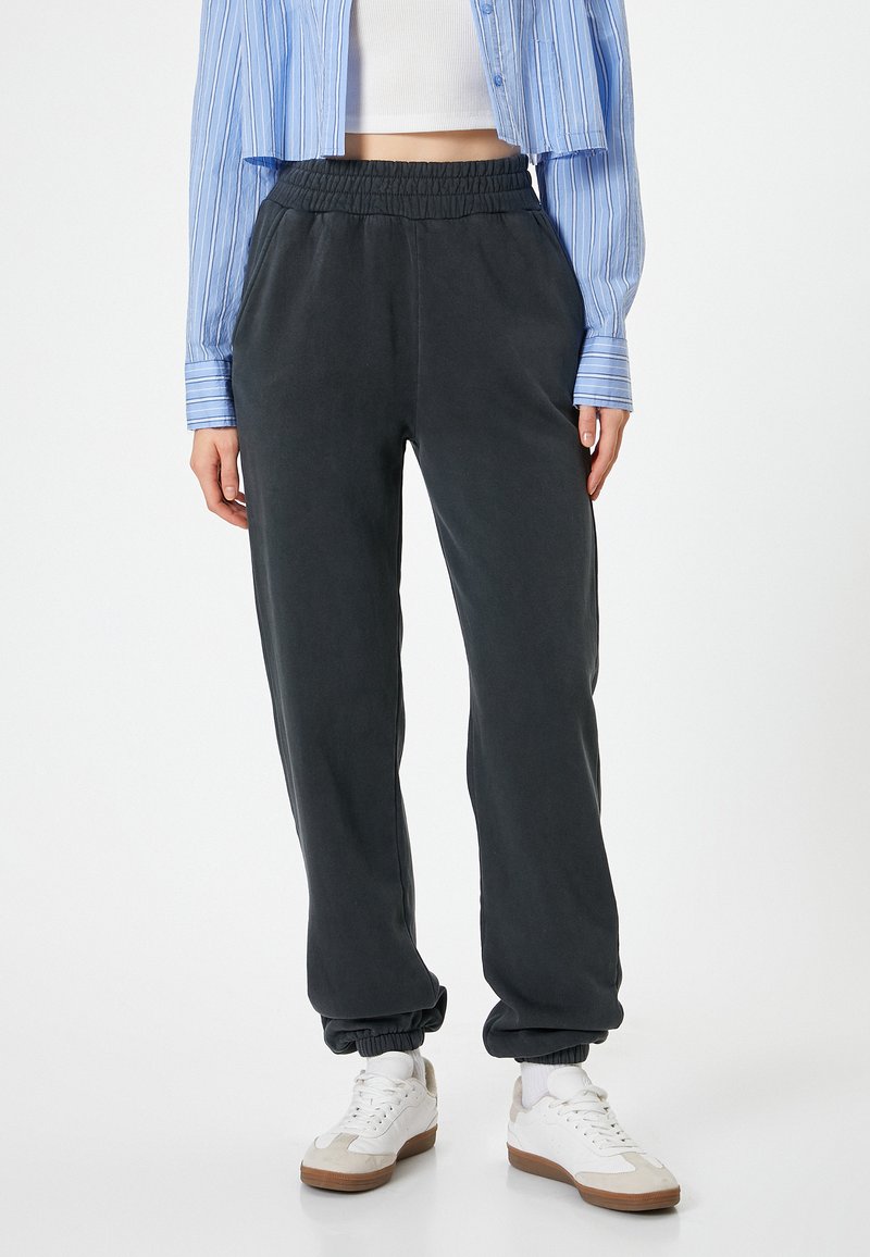 Dark grey jogger pants with an elastic waistband and cuffed ankles, made from a smooth fabric. Worn with a cropped blue striped shirt and sneakers.