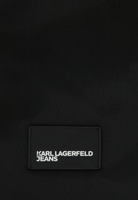 Črn tekstilni oznak z napisom "KARL LAGERFELD JEANS" v belem besedilu; gladka tekstura z pravokotno obliko in obrobljenimi robovi.