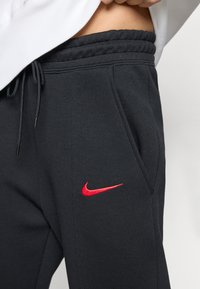 Sorte joggebukser med elastisk liv, snor, sidelommer og rødt Nike-logo på venstre lår. Myk, glatt stofftekstur.