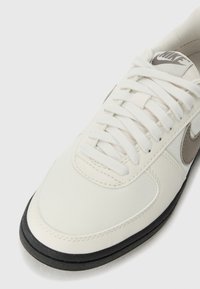 White Nike sneaker featuring a canvas upper, thick black rubber sole, round toe, and laces. Includes a subtle grey accent logo.