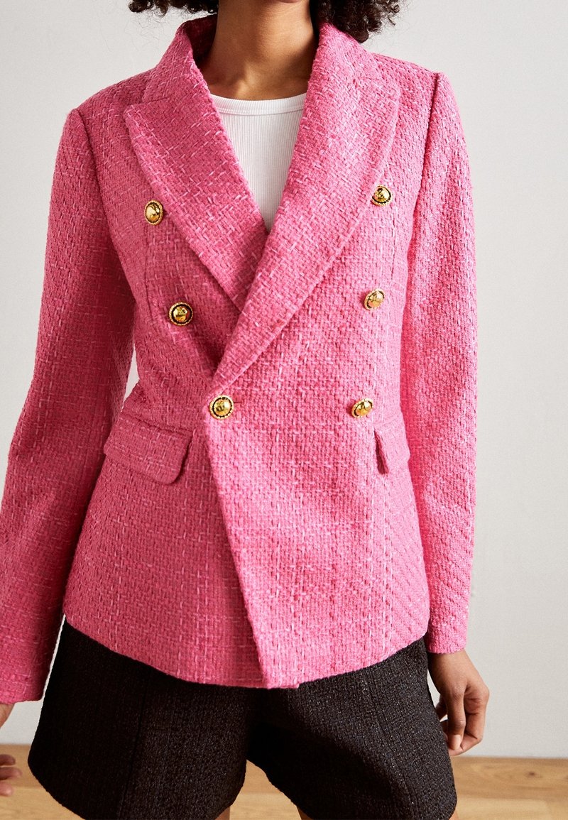 Blazer doppiopetto in tweed rosa con bottoni dorati, tessuto strutturato, rever a incavo e due tasche frontali. Indossato sopra una maglietta bianca.