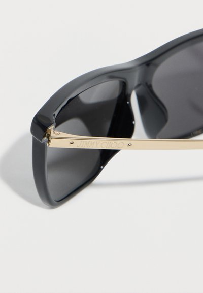 Μαύρα γυαλιά ηλίου με χρυσά μεταλλικά πτυσσόμενα μέρη, χαραγμένα με το λογότυπο "JIMMY CHOO" σε λευκό φόντο.