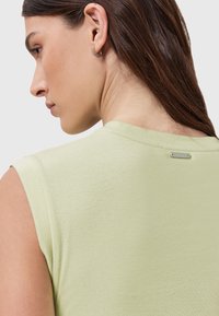 Lys grøn ærmeløs top i blødt stof, med en rund halsudskæring og et lille metalmærke på ryggen. Minimalistisk design.