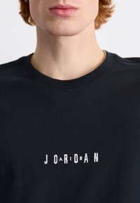 Maglietta nera in cotone con scollo rotondo. Presenta un design di testo ricamato bianco con la scritta "J O A R I D R A N" sul petto.