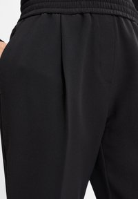 Zwarte stretchbroek met elastische tailleband en zijzakken, gedragen door een persoon met één hand gedeeltelijk in een zak.