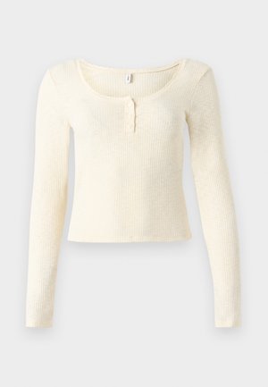 Világos krémszínű, hosszú ujjú bordás kötött felső; kerek nyakkivágással és háromgombos patenttal. Puha tapintású és testhez simuló szabású.